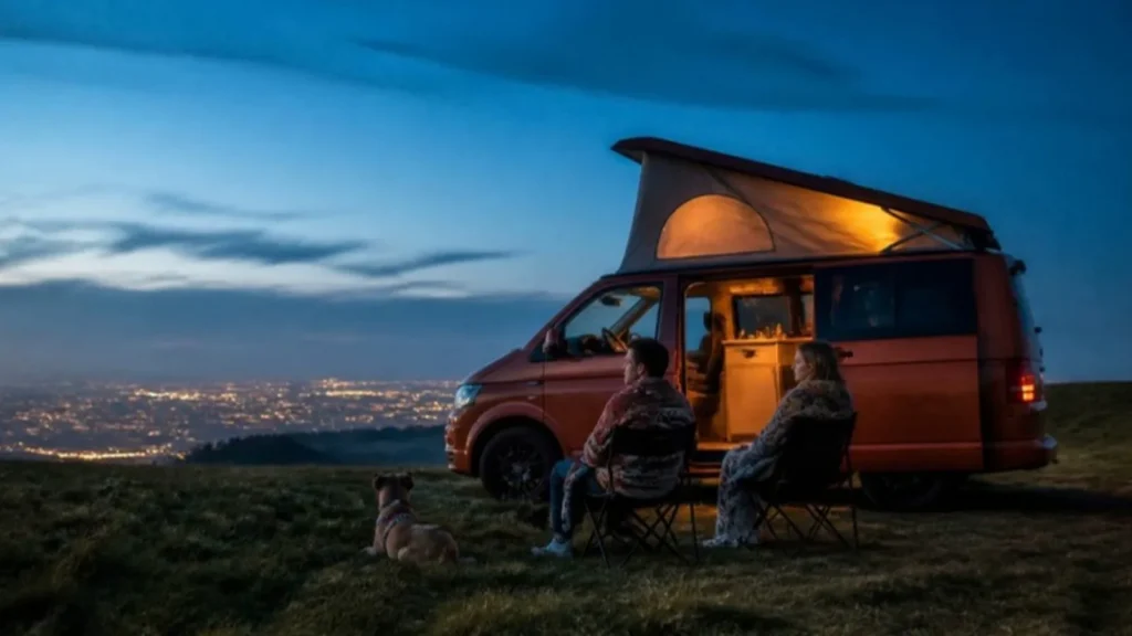 Couple and dog sitting by an orange pop top campervan on a hillside above city lights at dusk