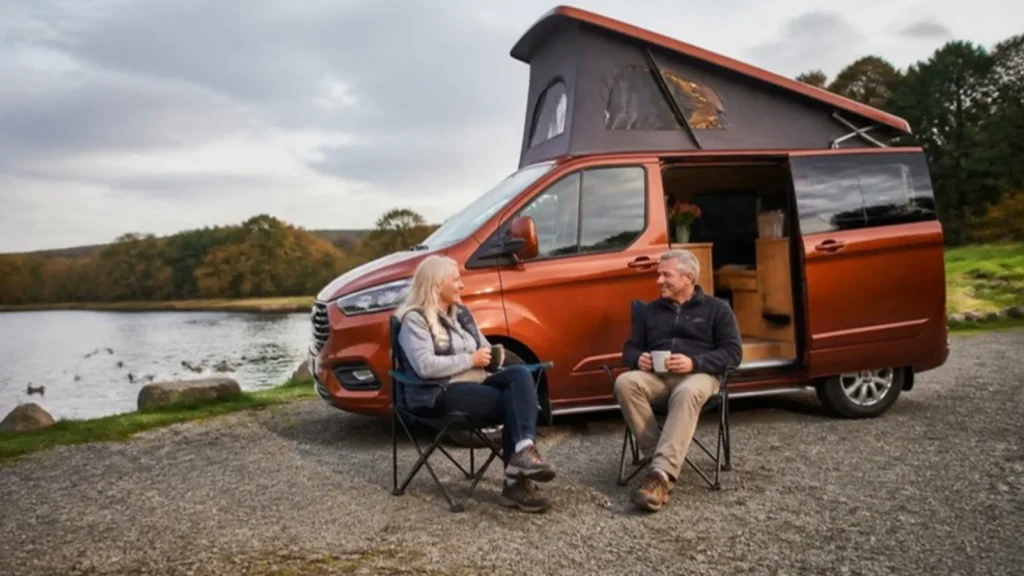 Couple with mugs in camping chairs beside an orange pop top campervan on a lake shore